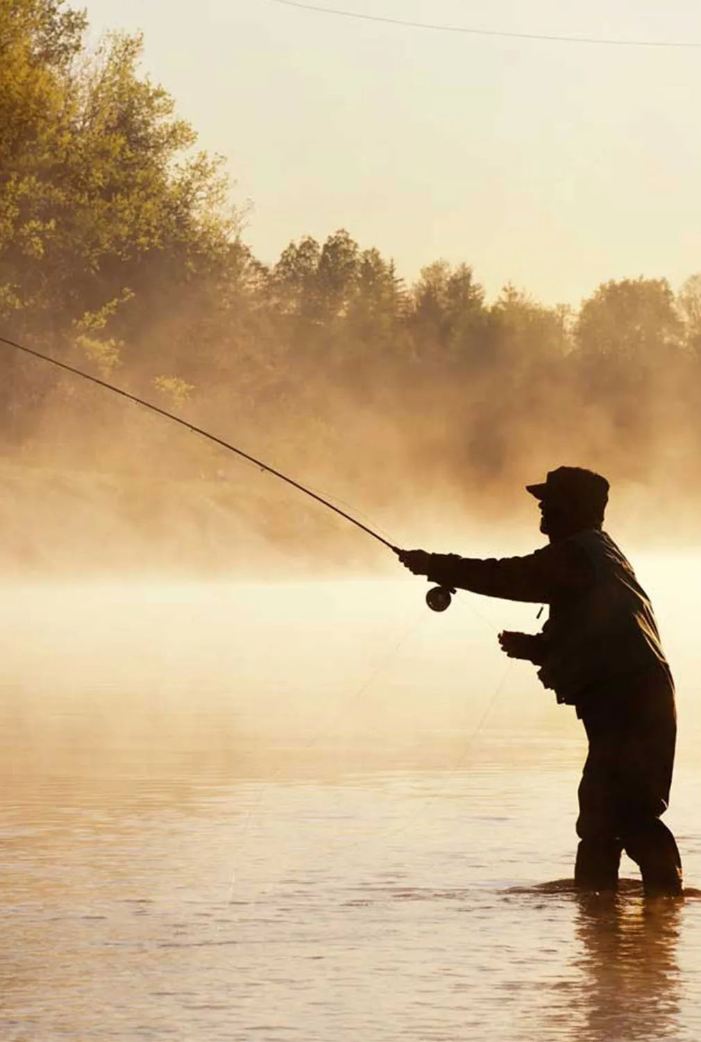 A man standing in a river up to his knees fly fishing during sunrise, creating a hazy back drop