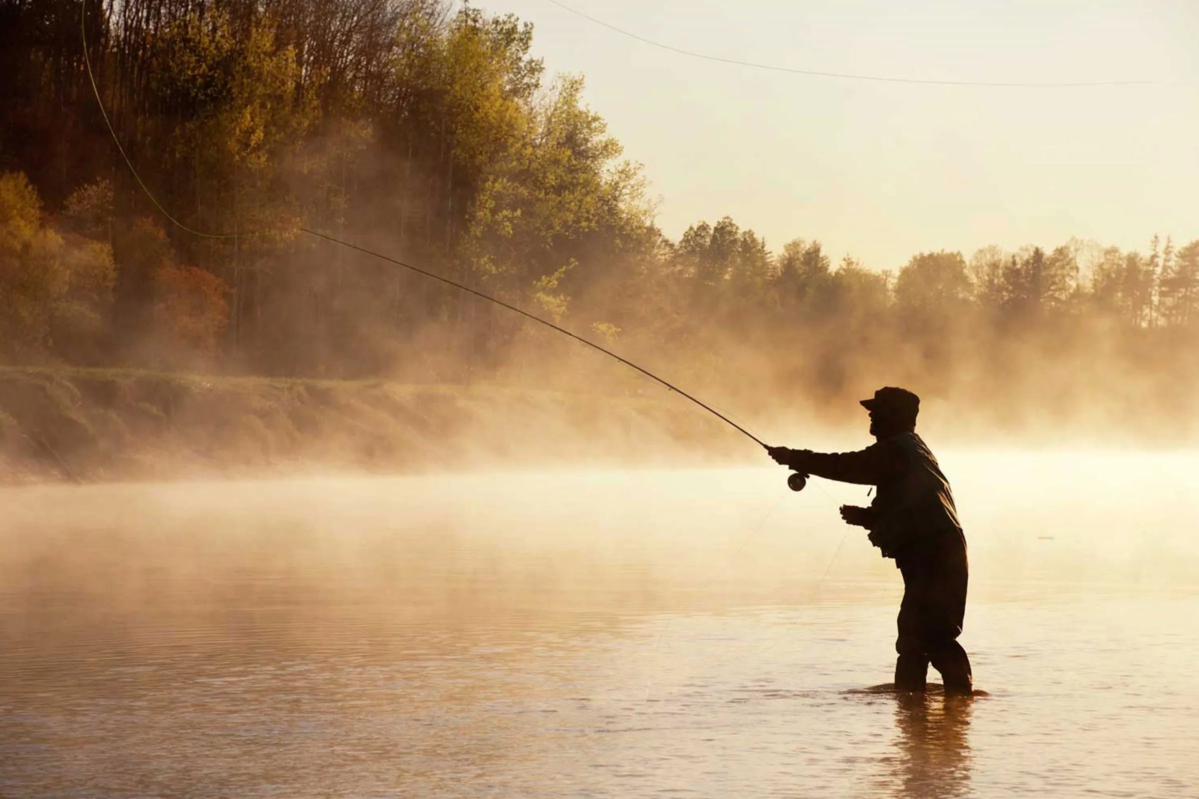 A man standing in a river up to his knees fly fishing during sunrise, creating a hazy back drop