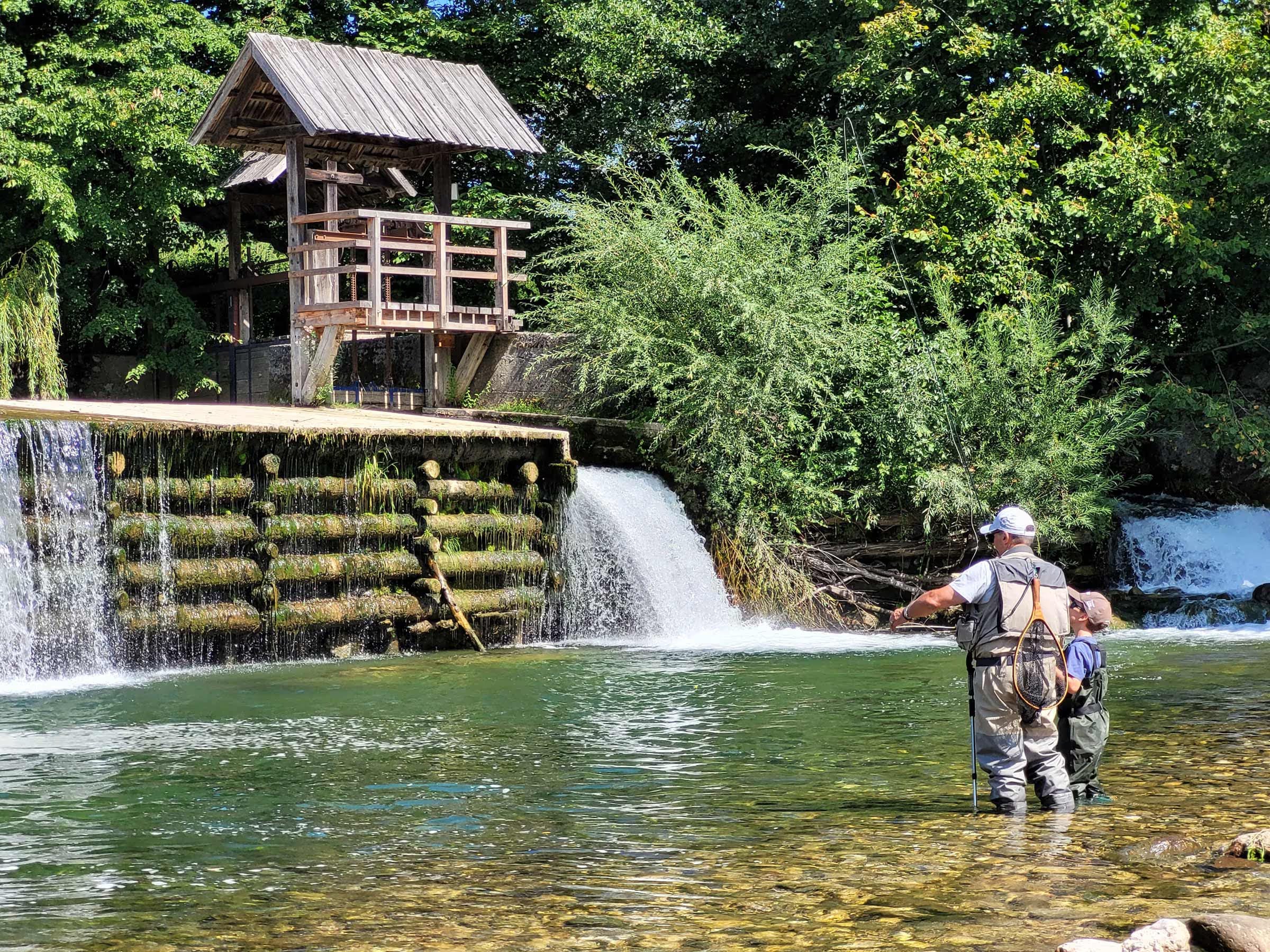 A farther and son fly fishing in river with a waterfall in the background